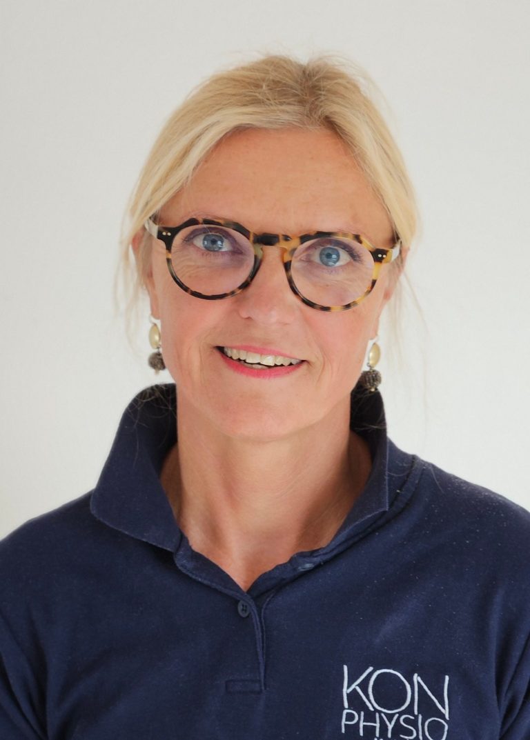 Physiotherapeutin Kathrin Ortinger Naujeck mit blonden Haaren und Brille trägt ein dunkles Polohemd mit KONPHYSIO Logo freundlich lächelnd.