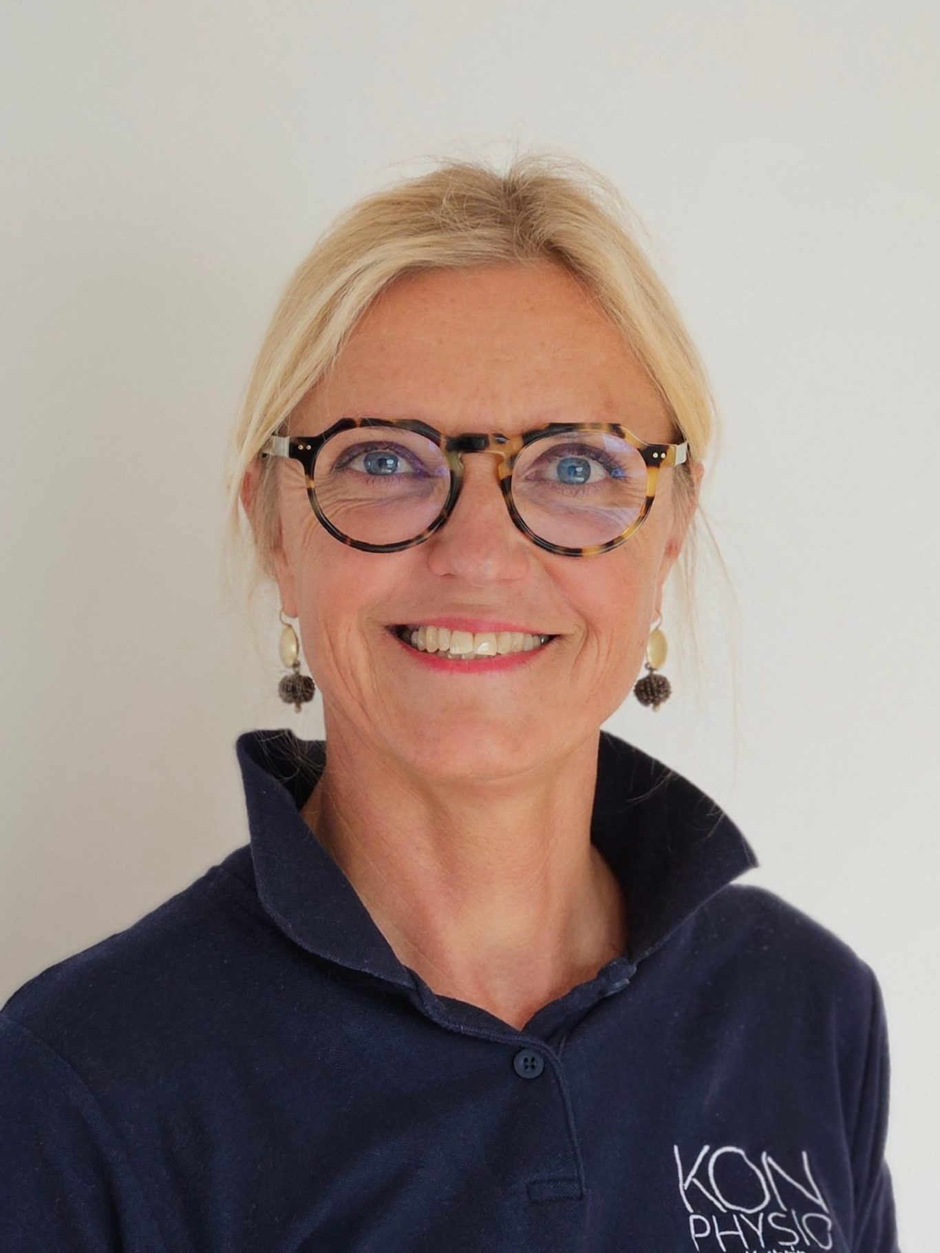 Lächelnde Physiotherapeutin Kathrin Ortinger Naujeck mit blonden Haaren, Brille und blauer Kleidung vor einem hellen Hintergrund.