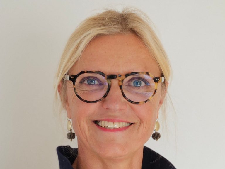 Physiotherapeutin Kathrin Ortinger Naujeck mit blonden Haaren, Brille und blauem Polo mit KONPHYSIO Logo, lächelt freundlich in die Kamera.