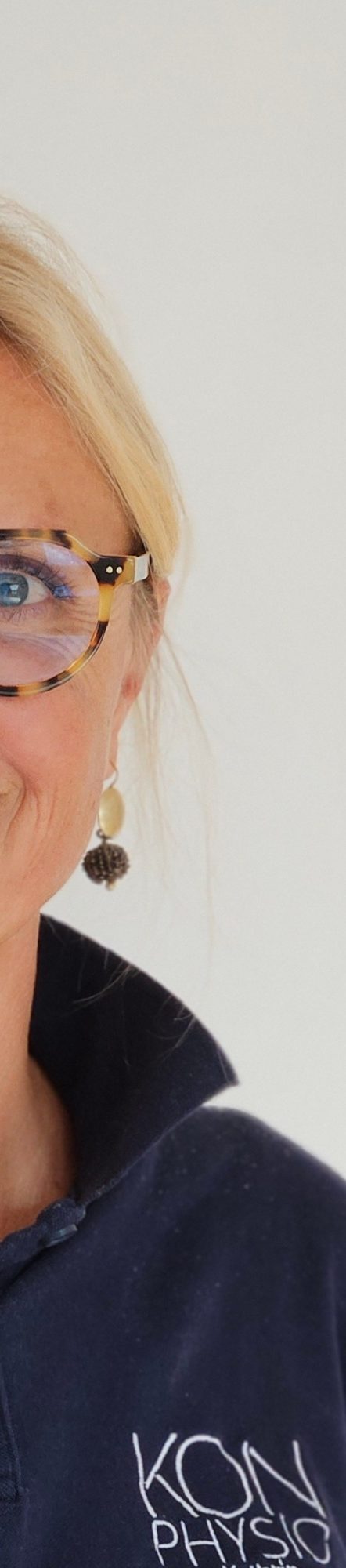 Physiotherapeutin Kathrin Ortinger Naujeck mit blonden Haaren, Brille und blauem Polo mit KONPHYSIO Logo, lächelt freundlich in die Kamera.