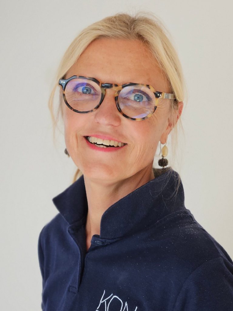Physiotherapeutin Kathrin Ortinger Naujeck mit blonden Haaren, Brille und blauem Polo mit KONPHYSIO Logo, lächelt freundlich in die Kamera.