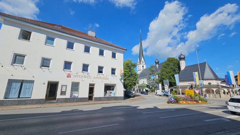 Zentrum Prien mit Haus Marktplatz 1, Kirche mit Fahnen und blühenden Blumenbeeten unter weißblauem Himmel.