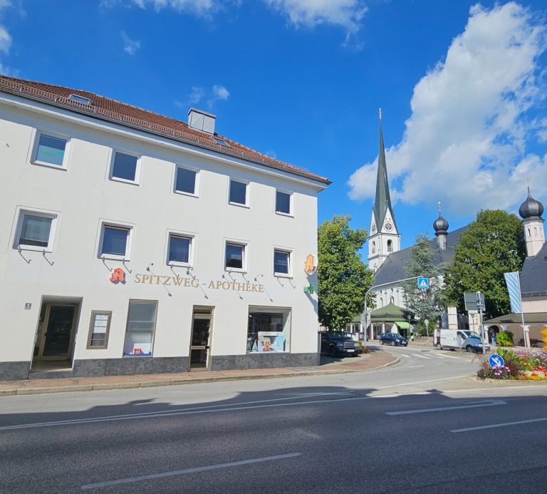 Blick auf das Haus der Spitzwegapotheke in Prien mit Kirche im Hintergrund unter weißblauem strahlenden Himmel
